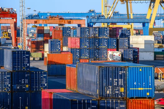 A lot of cargo containers stacked in shipping port under clear blue skies. Gantry cranes in background, ready to load and unload goods from ships.
