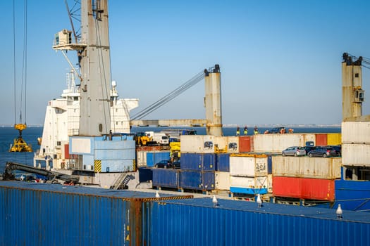 Cargo containers stacked with cars on top on bulk carrier in sea port on sunny day. Maritime logistics, transportation, commercial seaport, shipping containers in various colors, cargo handling, vessel deck cranes, clear blue sky, seagulls resting