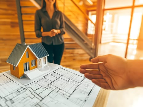 A real estate consultation scene with a miniature house model and blueprints on a table. A blurred woman stands in the background while a hand gestures, symbolizing property investment and home buying.
