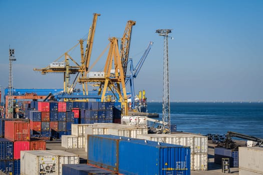 Maritime logistics and transportation scene at commercial seaport with stacked shipping containers, gantry and STS cranes, reach stackers in cargo terminal. International shipping and freight infrastructure, clear blue sky and sea waters.