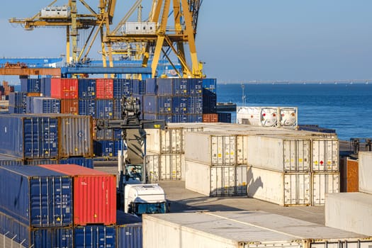 Cargo containers stacked on berth of sea port on sunny day. Maritime logistics, transportation, commercial seaport, global trade, shipping containers in various colors, cargo handling equipment, clear blue sky, ship-to-shore and gantry cranes.