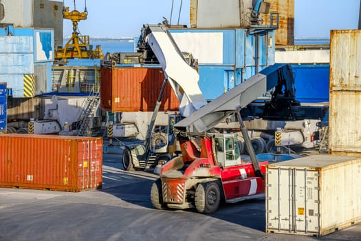 Two reach stackers and cranes transporting large cargo containers at shipping port. Bright sunlight, clear blue sky, stacked containers of various colors, shipping operations of sea cargo port.