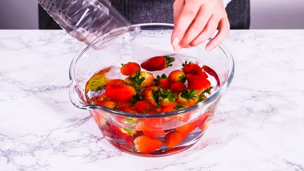 Ripe strawberries are submerged in water within a large glass mixing bowl, a step in washing the fruit to ensure cleanliness and longevity before storage or consumption.