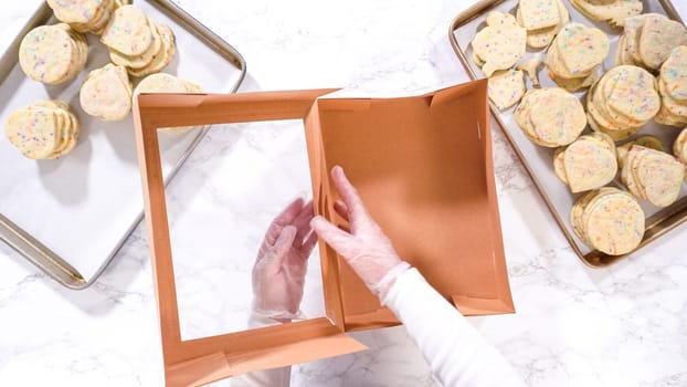 Flat lay. With precision, the woman is carefully arranging the sugar cookies, filled with dough-mixed sprinkles, into a rustic brown paper box.