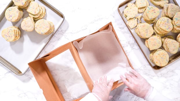 Flat lay. With precision, the woman is carefully arranging the sugar cookies, filled with dough-mixed sprinkles, into a rustic brown paper box.
