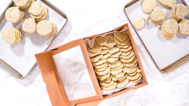 Flat lay. With precision, the woman is carefully arranging the sugar cookies, filled with dough-mixed sprinkles, into a rustic brown paper box.