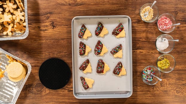 Buttery cookies half-dipped in chocolate, sprinkled with festive red, green, and white decorations, laid on white parchment to set.