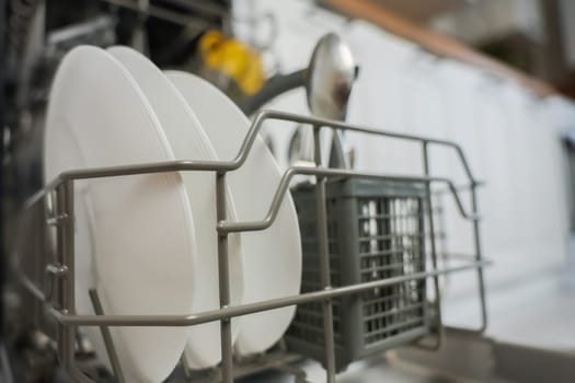 Close-up of open dishwasher department with clean plates and cutlery against kitchen background: white cabinets and wooden countertop out of focus. The concept of washing dishes, cleanliness, alleviating household chores. High quality photo