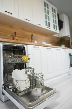 Vertical photo of open dishwasher department with clean plates and cutlery against light kitchen background with white cabinets and wooden countertop and panel. The concept of washing dishes, cleanliness, alleviating household chores. High quality photo