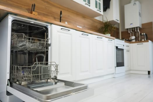 Empty dishwasher with two compartments in gray tones stands with the door open, ready to load, against the background of a white and wooden kitchen in out-of-focus. High quality photo