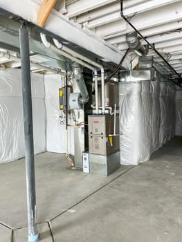 Castle Pines, Colorado USA - January 18, 2023-Image of the HVAC system and utility setup in the basement of a new construction single-family home. Visible are a furnace, ductwork, Aprilaire humidifier, and tankless water heater. The basement features white vapor barrier insulation, exposed ceiling beams, and an unfinished concrete floor. A metal support beam is also present, showcasing the structure of the home.