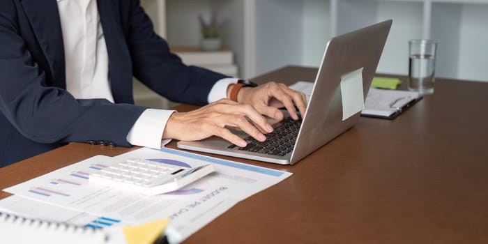 An elderly businessman focused on typing financial data on a laptop, with a calculator and reports spread across the desk in a modern office setting.