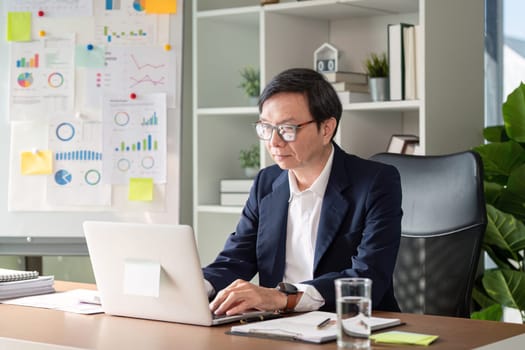 Focused elderly businessman working on data analysis using a laptop, surrounded by charts and graphs in a contemporary office setting.