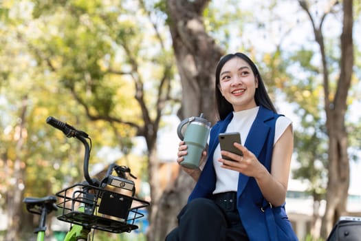 A cheerful businesswoman enjoys a moment in a sustainable park, holding her reusable drink and smartphone beside her green bicycle.