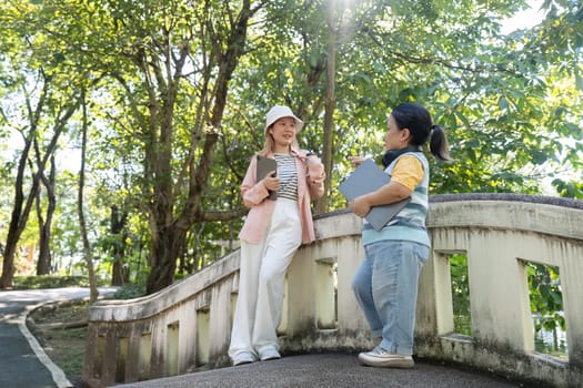 Two friends, one with achondroplasia, engage in a friendly conversation on a picturesque bridge surrounded by nature, with tablets in hand.