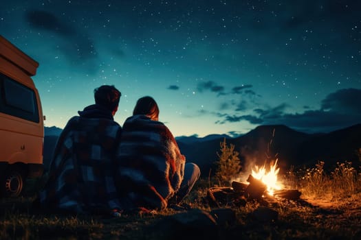 A couple sits close by a warm bonfire, wrapped in cozy blankets, as they gaze at the starry night sky. Their camper van stands nearby, creating a perfect outdoor adventure atmosphere.