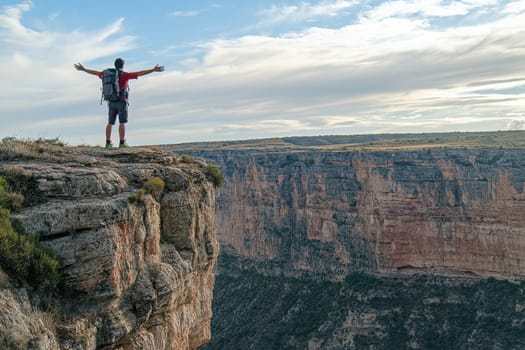 A backpacker stands at the edge of a dramatic cliff, arms wide open, embracing the stunning natural landscape surrounded by mountains and valleys.