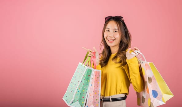 A beautiful Asian woman, a portrait of happiness, smiles joyfully while holding multicolored shopping bags. She stands elegantly with sunglasses in a studio shot on a pink background with copy space.