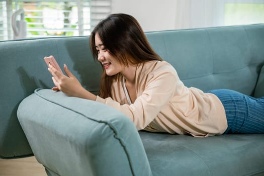 Smiling attractive woman relaxed reading text message on smartphone, Asian young female using smart mobile phone lying on sofa in living room room at home, gadget online application making internet