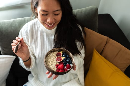 Happy young Asian woman smiling eating bowl of healthy breakfast oats with fruit sitting on the sofa. Healthy lifestyle concept.