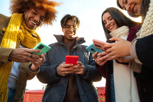 Smiling group of multiracial friends looking at their mobile phone on winter day. Addicted to social media and internet. Technology and addiction concept.
