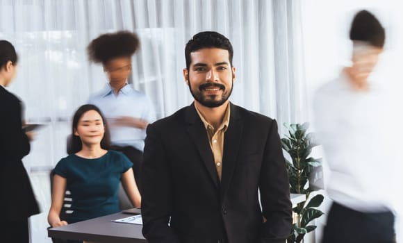 Businessman portrait poses confidently with diverse coworkers in busy meeting room in motion blurred background. Multicultural team works together for business success. Concord