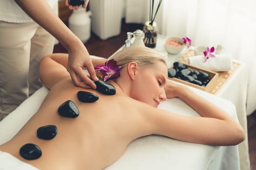 Hot stone massage at spa salon in luxury resort with day light serenity ambient, blissful woman customer enjoying spa basalt stone massage glide over body with soothing warmth. Quiescent