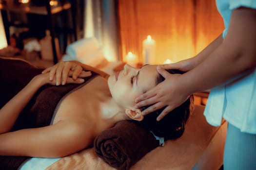 Caucasian woman enjoying relaxing anti-stress head massage and pampering facial beauty skin recreation leisure in warm candle lighting ambient salon spa in luxury resort or hotel. Quiescent