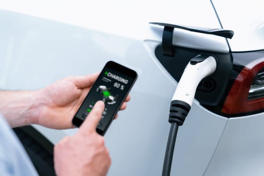 Modern eco-friendly man recharging electric vehicle from EV charging station, using Innovative EV technology utilization for tracking energy usage to optimize battery charging on smartphone.Synchronos