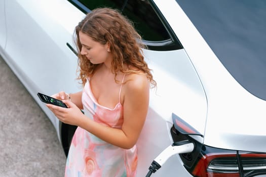 Aerial top view eco-friendly woman recharge electric vehicle from EV charging station, using EV technology utilization for tracking energy usage to optimize battery charging on smartphone. Synchronos