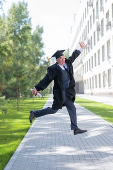 Old happy man in graduation gown jumping outdoors and holding diploma. Vertical