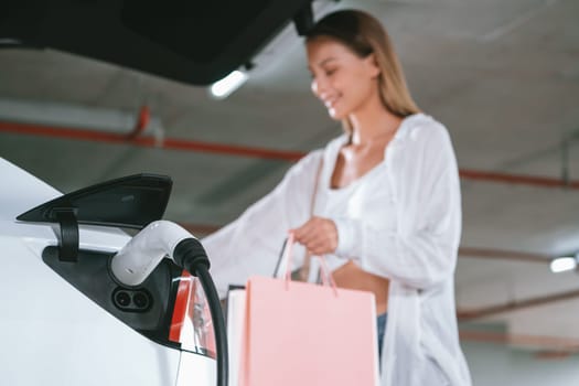 Young woman travel with EV electric car to shopping center parking lot charging in downtown city showing urban sustainability lifestyle by green clean rechargeable energy of electric vehicle innards