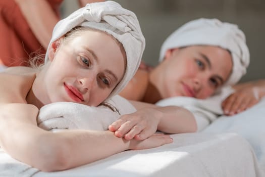 A couple of beautiful young woman with white towel lie on comfortable spa bed during receive relaxing body massage by professional masseur surrounded at spa salon. Gray background. Tranquility.