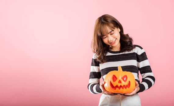 In a Halloween-themed studio shot on pink background, a joyful Asian woman holds model pumpkins, including a playful ghost-shaped one. Her festive costume enhances the Halloween experience.