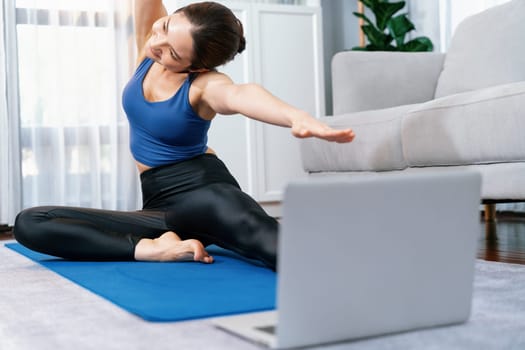 Focused laptop on the floor showing online exercise training video, while sporty athletic woman concentrate on warm-up and stretching routine in blurred background. Vigorous