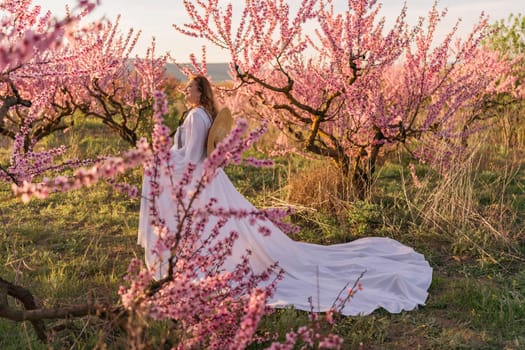 Woman blooming peach orchard. Against the backdrop of a picturesque peach orchard, a woman in a long white dress and hat enjoys a peaceful walk in the park, surrounded by the beauty of nature