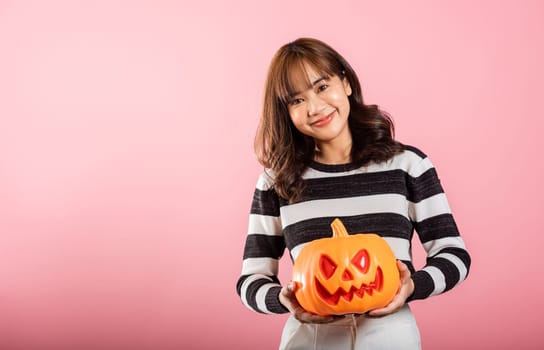 An Asian woman embraces the Halloween spirit in a studio shot on pink background, holding model pumpkins, including one in the shape of a ghost. Her cheerful costume adds to the fun.