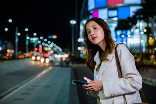 Close up of young Asian woman using mobile app device on smartphone to arrange taxi ride in city street. I Illuminated busy city traffic scene during rush hour with traffic congestion at night.