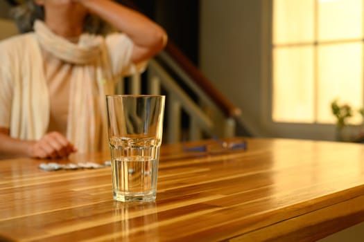 Close up of a glass of water on wooden table with blurred background of senior woman taking medication.