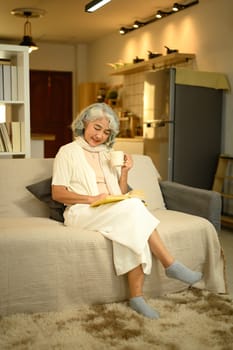 Serene mature woman drinking hot tea while reading book on sofa. Relaxation and leisure at home concept.