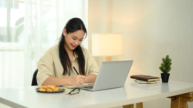 Smiling young woman writing in a notebook while working on her laptop.
