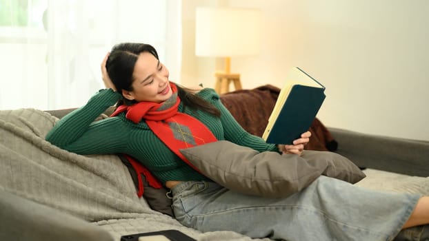 Cheerful young woman in green sweater and red scarf reading a book on couch.