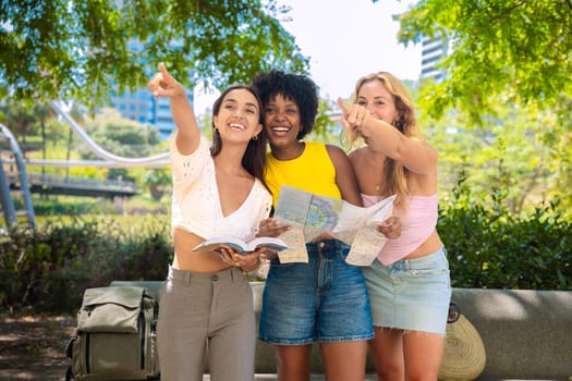 Three cheerful young women stand outdoors, holding maps and a travel guide while pointing at something. They smile excitedly, enjoying their adventure in a sunny urban setting with trees and buildings