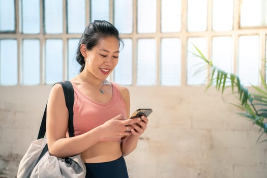 A cheerful woman wearing stylish activewear engages thoughtfully with her smartphone in a spacious and bright gym area