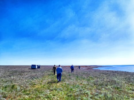 A group of individuals strolls across a sprawling grassland adjacent to a calm shoreline under a bright blue sky.