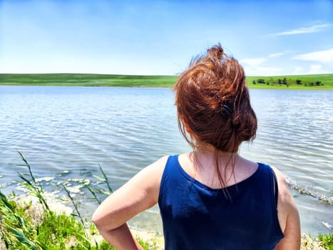 The woman stands by the water's edge, gazing at the peaceful lake and rolling hills under a clear blue sky.