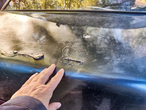 A hand touches the damaged area on a vehicle's hood while inspecting the paint and surface.