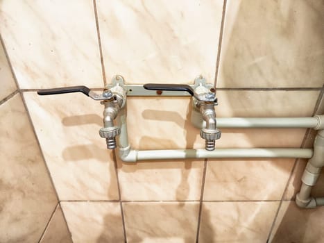 Two metallic plumbing taps are mounted on a tiled wall, connected by pipes, and ready for use in a utility space.