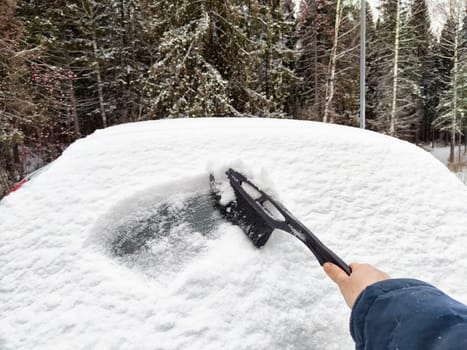 A person removes snow from the roof of a car using a brush on a cold winter day, preparing for a drive in snowy conditions.