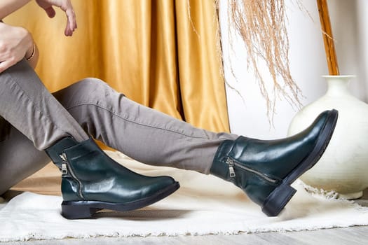 Model showcases modern green ankle boots while sitting on the floor in a minimalist setting featuring warm colors.